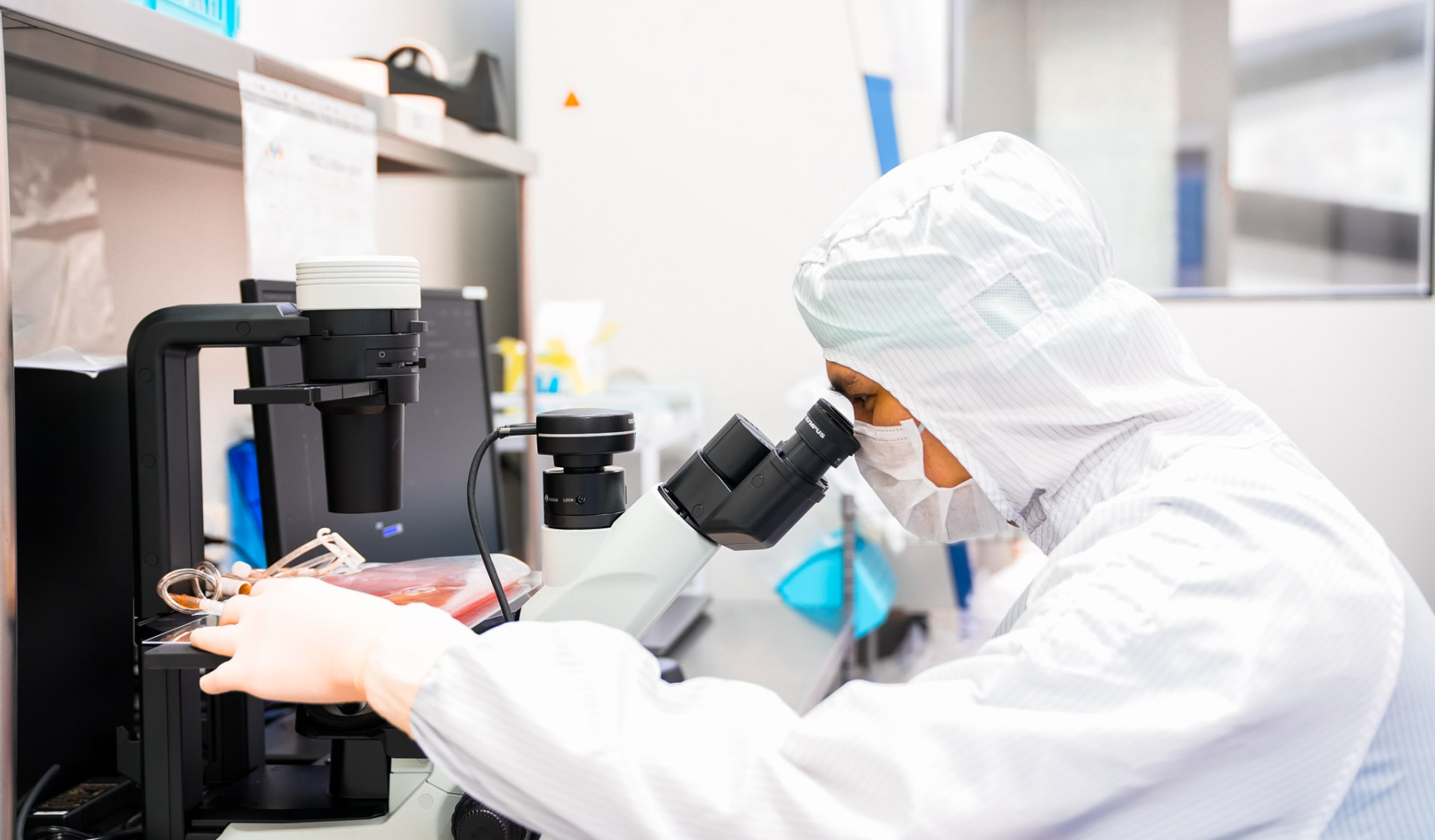 Laboratory scientist in full cleanroom suit examining a cell culture under a microscope